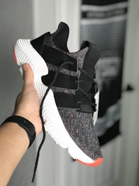 A hand holds a stylish sneaker with a black and white knit upper, featuring black straps and red accents. The shoe has a distinctive textured white midsole. There's a blurred background of a door and wall with unclear details.