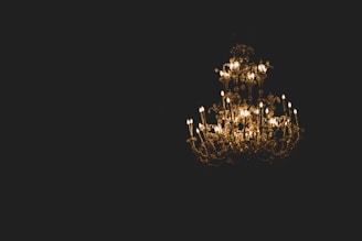 A large, ornate chandelier is suspended in a dark setting, featuring numerous glowing candle-like lights. The intricate metalwork and crystal decorations reflect warm light, creating a luxurious and elegant atmosphere.