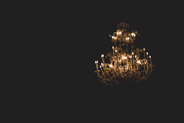 A large, ornate chandelier is suspended in a dark setting, featuring numerous glowing candle-like lights. The intricate metalwork and crystal decorations reflect warm light, creating a luxurious and elegant atmosphere.