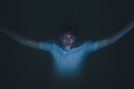 A person with outstretched arms appears submerged or floating in dark water, with a soft light illuminating their face and upper body. The scene conveys a sense of tranquility and isolation.