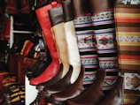 A collection of colorful and intricately designed boots are displayed, featuring a variety of leather textures and vibrant woven patterns. The boots range from solid red and brown leather to multicolored geometric designs, creating an eclectic and artisanal aesthetic.