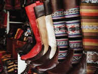 A collection of colorful and intricately designed boots are displayed, featuring a variety of leather textures and vibrant woven patterns. The boots range from solid red and brown leather to multicolored geometric designs, creating an eclectic and artisanal aesthetic.