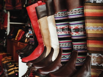 A collection of colorful and intricately designed boots are displayed, featuring a variety of leather textures and vibrant woven patterns. The boots range from solid red and brown leather to multicolored geometric designs, creating an eclectic and artisanal aesthetic.