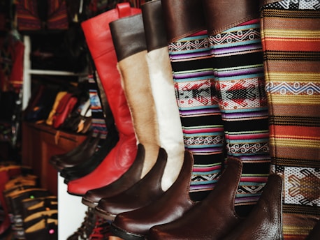 A collection of colorful and intricately designed boots are displayed, featuring a variety of leather textures and vibrant woven patterns. The boots range from solid red and brown leather to multicolored geometric designs, creating an eclectic and artisanal aesthetic.
