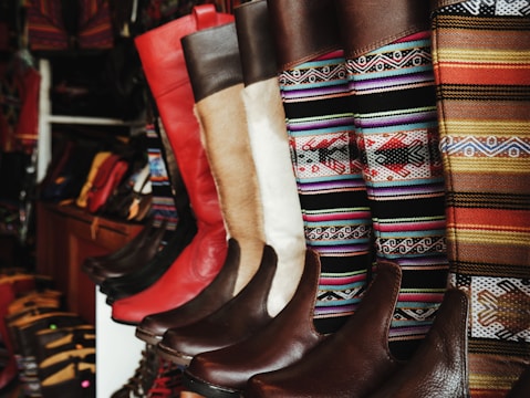 A collection of colorful and intricately designed boots are displayed, featuring a variety of leather textures and vibrant woven patterns. The boots range from solid red and brown leather to multicolored geometric designs, creating an eclectic and artisanal aesthetic.