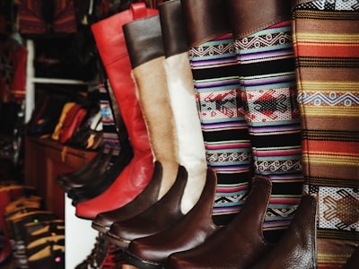 A collection of colorful and intricately designed boots are displayed, featuring a variety of leather textures and vibrant woven patterns. The boots range from solid red and brown leather to multicolored geometric designs, creating an eclectic and artisanal aesthetic.