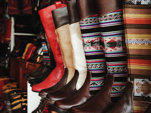 A collection of colorful and intricately designed boots are displayed, featuring a variety of leather textures and vibrant woven patterns. The boots range from solid red and brown leather to multicolored geometric designs, creating an eclectic and artisanal aesthetic.