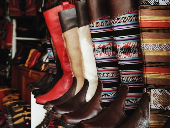A collection of colorful and intricately designed boots are displayed, featuring a variety of leather textures and vibrant woven patterns. The boots range from solid red and brown leather to multicolored geometric designs, creating an eclectic and artisanal aesthetic.