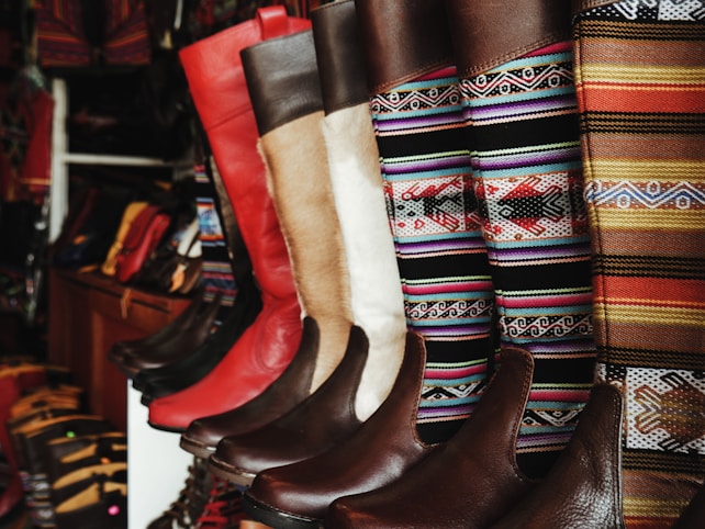 A collection of colorful and intricately designed boots are displayed, featuring a variety of leather textures and vibrant woven patterns. The boots range from solid red and brown leather to multicolored geometric designs, creating an eclectic and artisanal aesthetic.