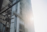 A high-rise building under construction is partially covered with scaffolding and netting. Sunlight creates a hazy effect, casting a soft glow on the structure. A utility pole with numerous electrical wires stretches from the foreground, enhancing the urban setting.