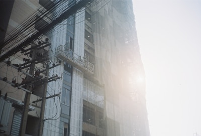 A high-rise building under construction is partially covered with scaffolding and netting. Sunlight creates a hazy effect, casting a soft glow on the structure. A utility pole with numerous electrical wires stretches from the foreground, enhancing the urban setting.
