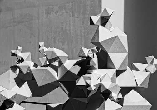 A geometric abstract sculpture composed of interconnected polyhedral shapes. The structure features a variety of angular forms, creating a complex and visually interesting pattern. The sculpture casts sharp shadows on a plain background, emphasizing its three-dimensional nature.