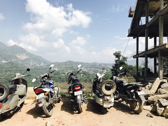 Multiple motorcycles and scooters are parked on a dirt patch near a partially constructed building. In the background, a scenic landscape of hills and a partly cloudy sky is visible. Rocks and building materials are scattered near the building.