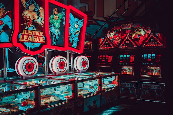 Brightly lit arcade machines with superhero-themed graphics featuring popular characters. The machines are adorned with colorful lights and feature comic book imagery, creating a vibrant and playful atmosphere.