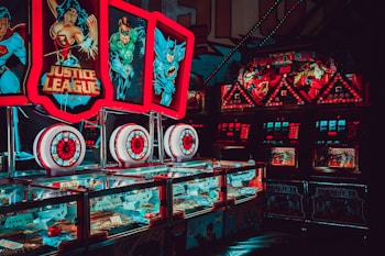 Brightly lit arcade machines with superhero-themed graphics featuring popular characters. The machines are adorned with colorful lights and feature comic book imagery, creating a vibrant and playful atmosphere.