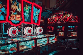 Brightly lit arcade machines with superhero-themed graphics featuring popular characters. The machines are adorned with colorful lights and feature comic book imagery, creating a vibrant and playful atmosphere.