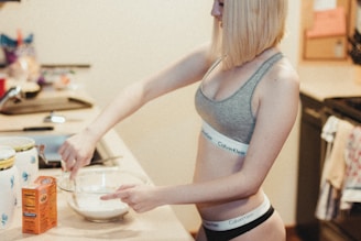 A person wearing a gray sports bra and black shorts is standing at a kitchen counter, mixing ingredients in a glass bowl. There is a box of baking soda on the counter, along with a container with a blue floral design. The kitchen appears to be well-lit, and various kitchen items and a dish towel are visible in the background.