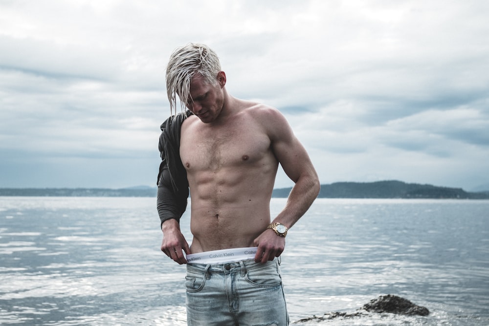 topless man wearing gray denim bottoms standing on shore at daytime