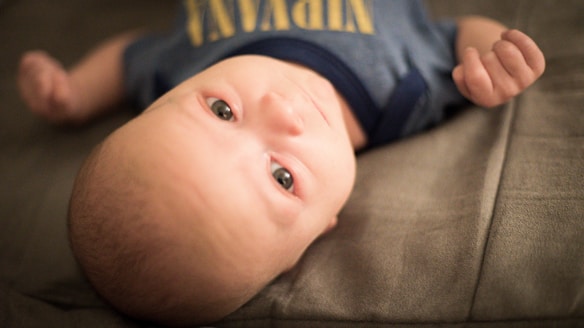A baby lies on a soft, neutral-colored surface, wearing a blue outfit with yellow lettering. The baby stares upwards with wide eyes and slightly clenched fists, conveying a sense of curiosity or alertness.