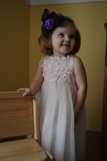 A young child with short hair is wearing a white dress adorned with floral patterns. The child also has a large headband with a purple flower and black bow. The background features a yellow wall and a wooden chair is partially visible.