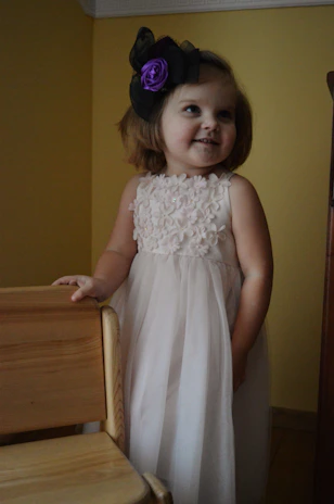 A young child with short hair is wearing a white dress adorned with floral patterns. The child also has a large headband with a purple flower and black bow. The background features a yellow wall and a wooden chair is partially visible.