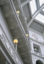 An ornate interior view of a building with a high ceiling featuring elaborate molding and architectural details. Several decorative fixtures and recessed panels are visible. Light fixtures with glowing bulbs are incorporated into the design. The structure is predominantly composed of light-colored materials, emphasizing its intricate carvings and majestic appearance.