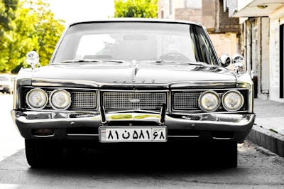 A well-preserved vintage car with a shiny chrome grille and headlights is parked on a street. The car's license plate displays characters in a non-Latin script. Trees and buildings form the background, illuminated by bright sunlight.