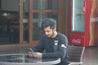 A person sitting at an outdoor table, looking at their smartphone with a focused expression. The setting appears to be casual, with a visible Coca-Cola vending machine in the background and glass doors leading to the interior of a building.
