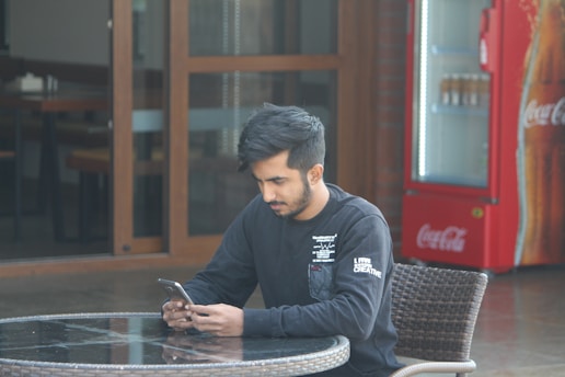 A person sitting at an outdoor table, looking at their smartphone with a focused expression. The setting appears to be casual, with a visible Coca-Cola vending machine in the background and glass doors leading to the interior of a building.