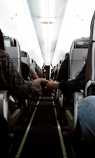 Interior view of an airplane cabin showing rows of seats with passengers seated. Two people sitting on opposite sides are holding hands across the aisle. Overhead compartments and a sign indicating 'SALIDA EXIT' are visible. The lighting is soft, suggesting a calm atmosphere.