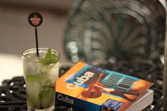 A refreshing cocktail garnished with mint leaves in a glass with a Havana Club stirrer sits on a patterned metal table. Next to the glass is a book about Cuba featuring a colorful cover.