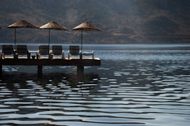 A serene lakeside scene with several lounge chairs and parasols on a wooden pier extending over calm water. The backdrop consists of mountains and a clear sky, creating a peaceful atmosphere.