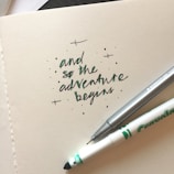 A close-up of a piece of paper featuring the handwritten phrase 'and so the adventure begins' in black and green ink. The paper is accompanied by a gray Staedtler pen and a white Crayola marker, both positioned diagonally across the text. Small decorative dots and crosses are scattered around the writing.