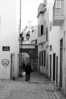 A black and white photograph of a narrow cobblestone street flanked by white buildings with shuttered windows. An archway leads further into the narrow passage, adorned with street lamps. A lone man walks down the street, casting a long shadow. A street sign with Arabic script and the name 'Rue du Tribunal' is visible, along with some graffiti on the wall.