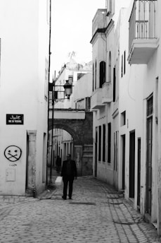 A black and white photograph of a narrow cobblestone street flanked by white buildings with shuttered windows. An archway leads further into the narrow passage, adorned with street lamps. A lone man walks down the street, casting a long shadow. A street sign with Arabic script and the name 'Rue du Tribunal' is visible, along with some graffiti on the wall.