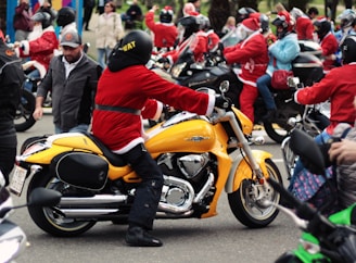 A group of people dressed in Santa outfits are riding motorcycles on a street, with one riding a prominent yellow motorcycle in the foreground. The scene suggests a festive or parade-like atmosphere with various models of bikes and people interacting.