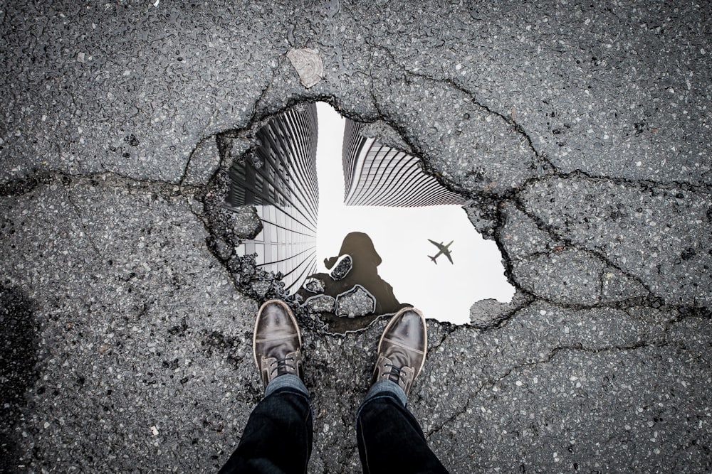 Person standing in front of a puddle on pavement, looking down at the puddle. There are reflections of city buildings and an airplane flying overhead in the puddle
