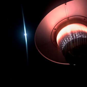 A glowing outdoor patio heater with a circular design and a prominent orange light emanating from its mesh-covered element. The heater is positioned against a dark background, with the bright light creating a dramatic contrast.