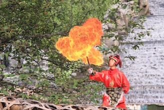 A performer dressed in a bright red traditional outfit is breathing fire, creating a large flame in front of lush green foliage. The intricate costume includes a headpiece with red ornaments. The setting appears to be outdoors, with trees and a textured stone wall in the background.