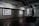 A dimly lit art gallery features several framed art pieces mounted on a textured concrete wall. There is a black display stand in the center of the room with a curved glass top, possibly showcasing an item underneath. The lighting is focused on the walls, highlighting the artwork while casting the rest of the room in shadows.