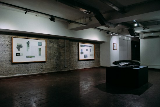 A dimly lit art gallery features several framed art pieces mounted on a textured concrete wall. There is a black display stand in the center of the room with a curved glass top, possibly showcasing an item underneath. The lighting is focused on the walls, highlighting the artwork while casting the rest of the room in shadows.