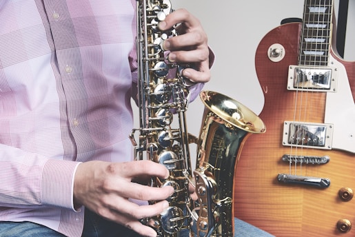 A person wearing a plaid shirt is holding and playing a saxophone. Beside the saxophone, an electric guitar in a sunburst finish can be seen, resting upright. The focus is on the hands and musical instruments.