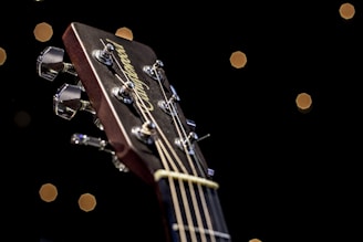 A close-up view of a guitar headstock, focusing on the tuning pegs and strings. The background is dark with small, softly illuminated circular lights resembling a bokeh effect.