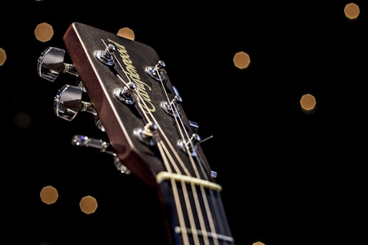 A close-up view of a guitar headstock, focusing on the tuning pegs and strings. The background is dark with small, softly illuminated circular lights resembling a bokeh effect.