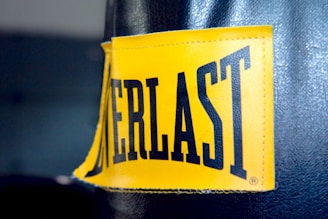 A close-up view of a yellow rectangular label with black stitching that has the word 'EVERLAST' printed in bold black capital letters. The label is attached to a dark-colored textured material, which appears to be part of a punching bag or similar sporting equipment.
