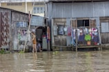 A person stands in floodwater in front of dilapidated houses with clothes hanging to dry. The water reaches up to the doorsteps, and there is a collection of household items visible. Another person is sitting inside the doorway of one of the houses.