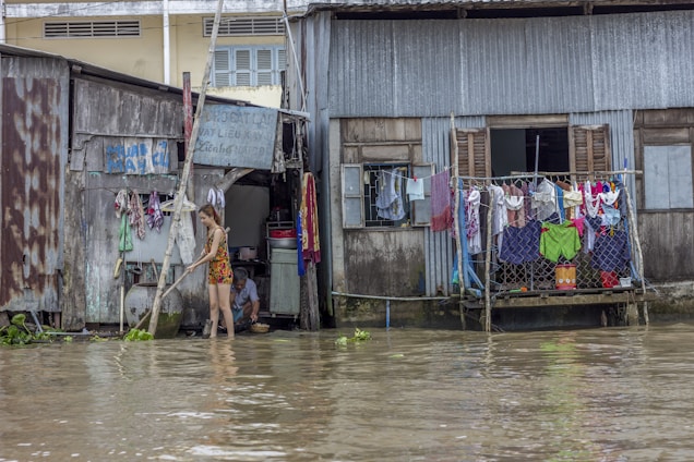 A person stands in floodwater in front of dilapidated houses with clothes hanging to dry. The water reaches up to the doorsteps, and there is a collection of household items visible. Another person is sitting inside the doorway of one of the houses.