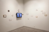 An art gallery wall displays several small frames, some with text and others with blank spaces, arranged asymmetrically. A television screen is mounted on the wall showing a man speaking.
