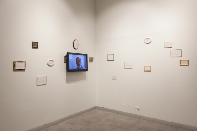 An art gallery wall displays several small frames, some with text and others with blank spaces, arranged asymmetrically. A television screen is mounted on the wall showing a man speaking.
