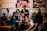 A group of people are gathered in a room with colorful abstract art on the walls. The central figure appears to be speaking or expressing a point, while others listen attentively. The setting suggests a casual yet focused discussion or meeting.